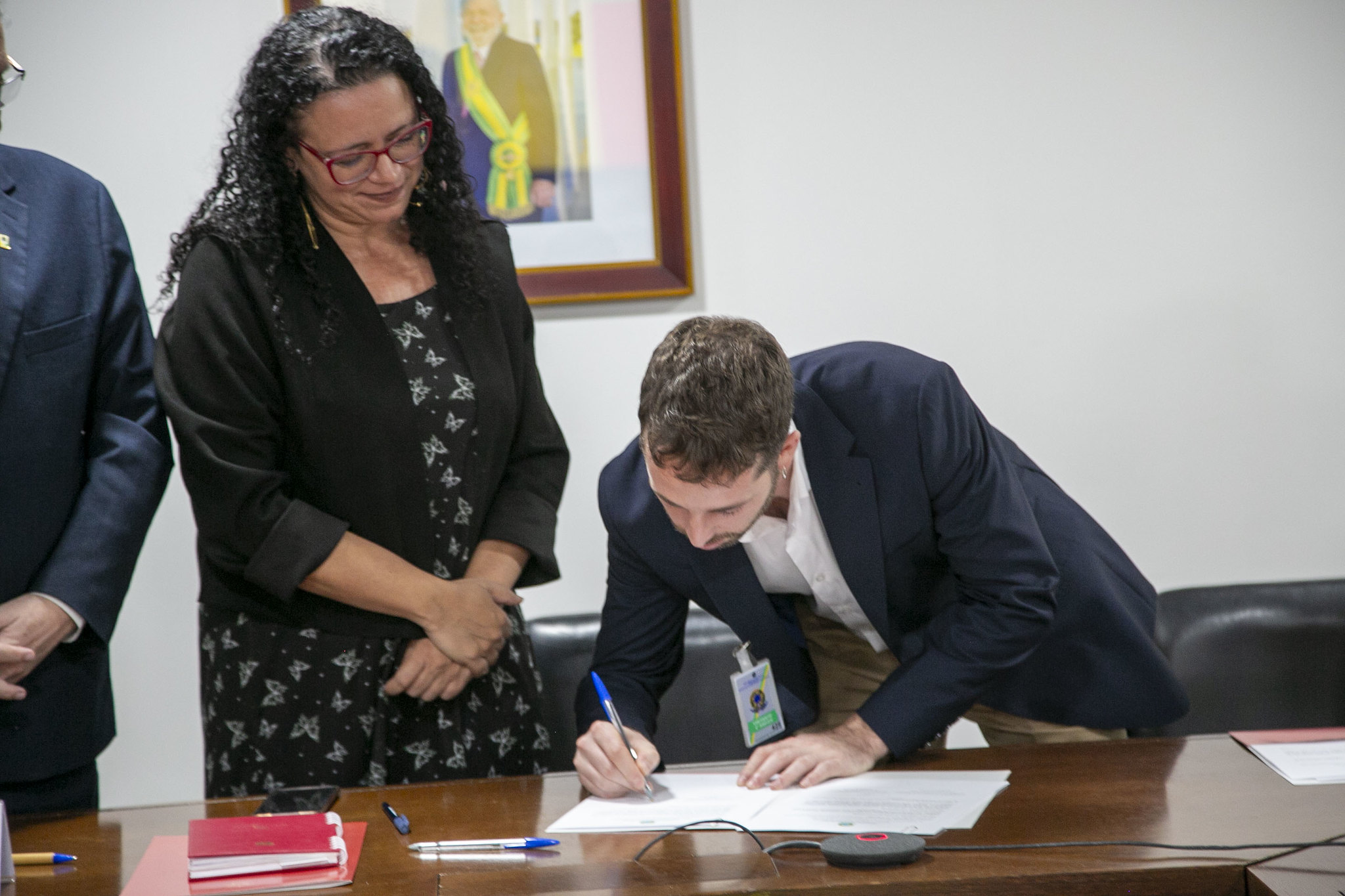 A man in a dark suit signs a document at a wooden desk while a woman in a black dress watches, with another person partially visible to the left. A framed portrait hangs on the wall behind them