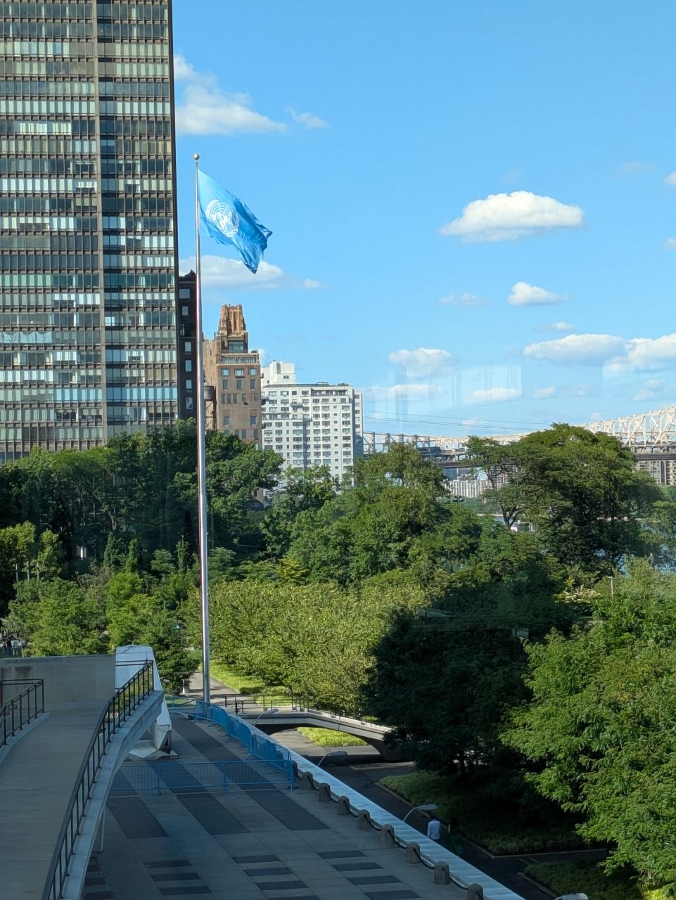The United Nations flag flies on a tall pole near the UN headquarters building, overlooking a green treeline with city buildings and a bridge in the background.