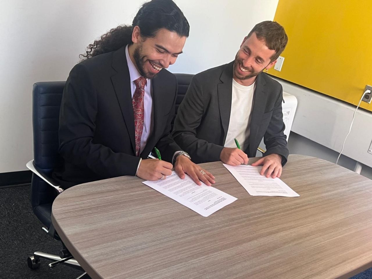 Two smiling men in suits sign documents at a wooden table in an office with a yellow wall in the background.