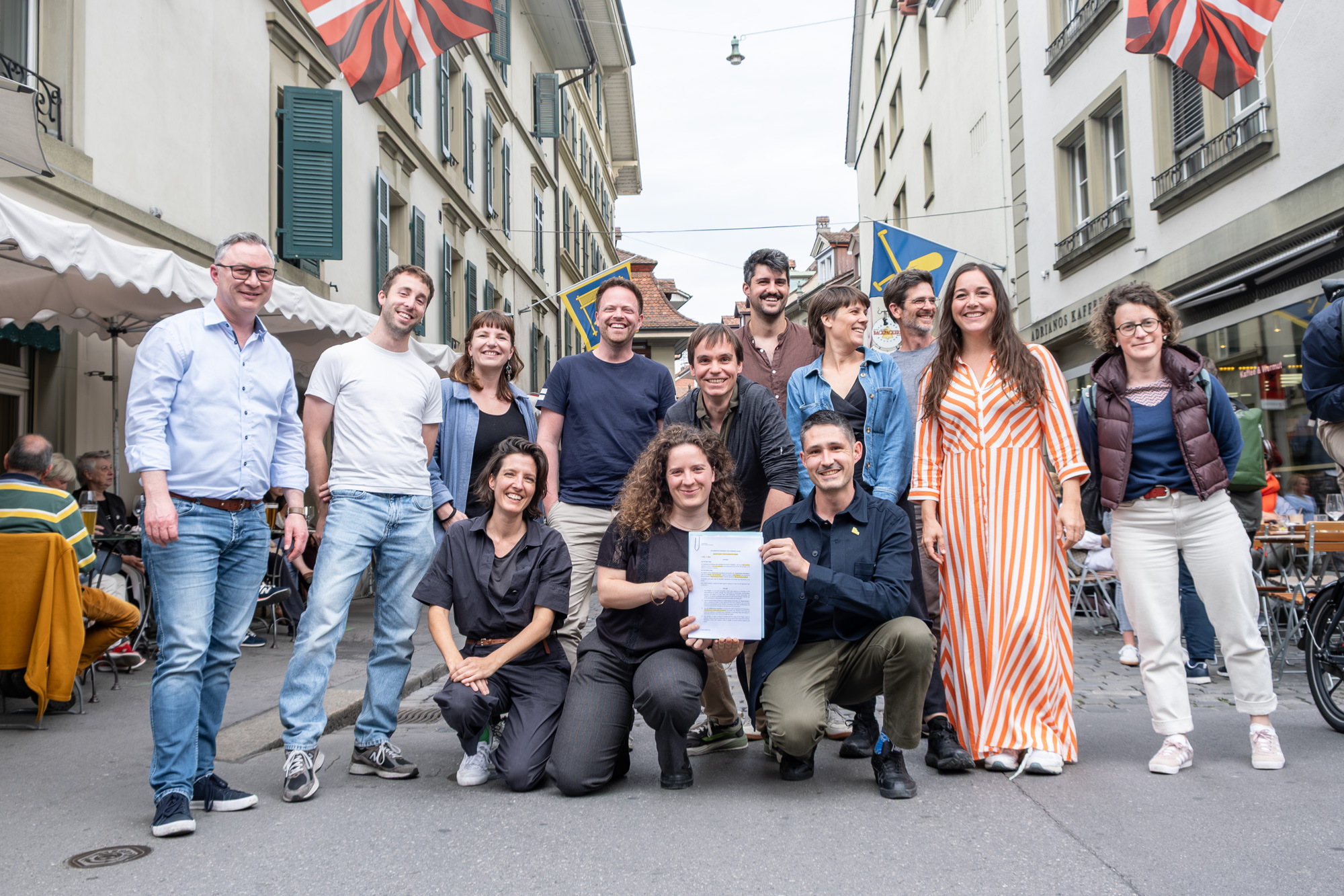 A group of people pose together on a European street lined with historic buildings and cafes, with three people crouched in front holding a document.